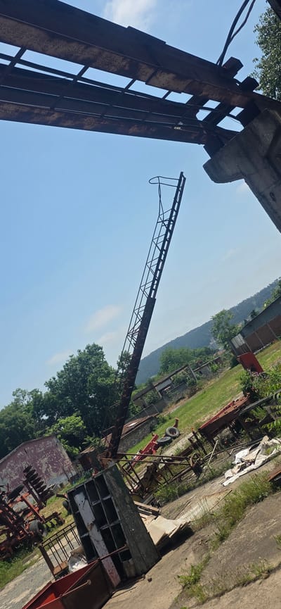 This image captures a wide outdoor scene at what appears to be a disused industrial or agricultural site in Karnobat, Bulgaria, under a clear, bright blue sky with faint wisps of clouds, suggesting a sunny day.

The foreground is dominated by large, heavily rusted metal structures. To the left, a massive, upright metal beam or support pillar, likely part of a larger industrial framework, stands prominently. Its intricate, lattice-like construction is visible, showing signs of significant age and weathering. Extending across the upper middle of the frame, a long, slender jib or boom of a crane, also made of rusted metal lattice, reaches horizontally into the sky, ending with a distinctive U-shaped or hook-like component.

The ground area below these structures is a mixture of overgrown grass, a paved or concrete path, and numerous pieces of discarded machinery and debris. Visible objects include a red-framed piece of equipment with two wheels, possibly part of a trailer or farm implement, and another rusty machine with multiple disc-shaped components, resembling a disc harrow. Various other metal fragments, wooden crates or shelving, and white tarps are scattered across the uneven ground. Lush green trees and vegetation are prominent in the middle ground, partially obscuring the distant landscape.

In the background, rolling hills or mountains are visible under the expansive sky, with a few distant buildings, potentially industrial or residential, interspersed among the trees. There is no human activity present in the scene, and no visible text can be discerned on any of the objects. The overall atmosphere conveys a sense of abandonment and the gradual decay of industrial or agricultural infrastructure.