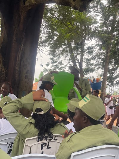The image depicts an outdoor gathering, possibly in Emure-Ekiti, Nigeria, as suggested by contextual information. A large tree trunk dominates the left side of the frame, with dense foliage overhead indicating a natural or park-like setting. Several individuals, primarily young adults, are present, many wearing matching olive green uniforms with green caps, suggestive of a uniformed youth service or a similar organization.

In the foreground, two individuals are seen from behind, also in the green uniform, seated on white plastic chairs. The chair in the lower right has "PL" and "in You" written on its back in black marker. The individual on the left is wearing a cap with a light yellow band and two green stripes.

In the mid-ground, a group of people are interacting. A woman with braided hair, wearing a white shirt and a green cap, is visible on the far left, looking towards the center. Next to her, a person in the green uniform is talking with another individual. A large, bright green plastic structure, possibly a decorative item or part of an event setup, is a prominent object in the middle of the scene. Behind it, a man in a green uniform is holding a bright blue bowl filled with various bottled beverages.

In the background, other individuals can be seen, some in white shirts and shorts, suggesting a diverse group of attendees or participants. The ground appears to be sandy or dirt. The sky is overcast, creating diffused lighting. The overall atmosphere suggests a community event or gathering.