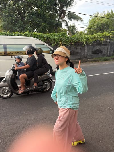 In Banjar, Indonesia, a young woman in a light blue long-sleeved shirt and pink pants, wearing a straw hat and glasses, gestures with her hand while standing on asphalt. She appears to be posing for a photo, with a hint of a smile. Behind her, a white van is parked, and to the left, lush green trees and a palm tree are visible. The scene suggests a warm, possibly sunny day.

Further in the background, three people are riding on a scooter. The person in the front is driving, wearing a blue shirt and a helmet. The middle person is wearing a black shirt and a helmet, and the person in the back is wearing a black shirt and a helmet. They are all facing forward, suggesting they are traveling. The scooter and the van are common sights in Indonesian transportation. The asphalt road and the dense greenery indicate an outdoor, likely tropical environment. The overall mood seems casual and relaxed, possibly during a tourist outing or a local journey. The image also features some visible text on the side of the van, but it's partially obscured.