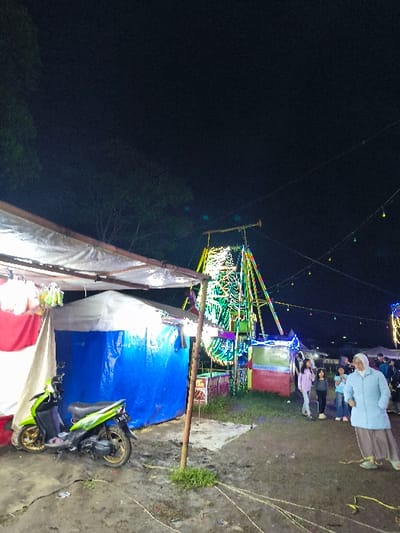 The image portrays an outdoor night scene in An Khe Ward, Vietnam, depicting a temporary fairground or market environment. In the left foreground, a light green scooter with gold-colored wheels is parked on muddy, uneven ground next to a white and green tent structure adorned with a red and white banner. Behind this, a second tent covered in a blue tarp is visible. A slender wooden pole stands upright from the ground near the center of the frame, where patches of grass and scattered yellow material, possibly tape or rope, are present.
In the midground, a prominent fairground ride, likely a Ferris wheel, is brightly illuminated with multi-colored LED lights in green, blue, yellow, and pink hues. Additional smaller, illuminated stalls or booths are situated near the base of this ride.
To the right, a woman wearing a light blue long-sleeved top, a white hijab, and light-colored pants stands in the foreground, facing towards the left. Behind her, a group of approximately five individuals, appearing to be children or young teenagers in casual clothing, are present. The background features a dark night sky with the silhouettes of trees visible on the left and strings of small, colored decorative lights strung across the upper midground.