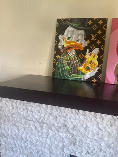 The image shows a painting of Scrooge McDuck wearing a black top hat and green plaid suit, holding a Bitcoin symbol in his gloved hands. The background of the painting features a Louis Vuitton-inspired pattern. The painting is placed on a dark brown shelf, which is mounted on a textured white wall. Another painting is partially visible to the right of the Scrooge McDuck painting. The image does not contain any information about the city of Ореховка, Russia, or its specific location within the city.