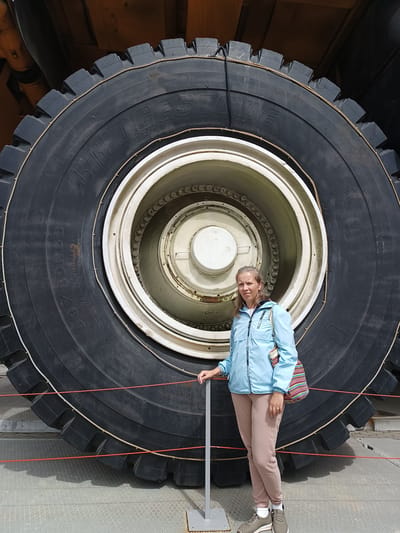 A woman stands in front of an enormous tire in the Museum of Military Technology in Verkhnyaya Pyshma, Russia. The tire is black with white lettering and has a thick tread pattern. The woman is wearing a light blue jacket, beige pants, and gray sneakers. She is holding onto a metal railing. The tire is part of a large piece of heavy machinery, likely a mining truck or construction vehicle. The background is dark and out of focus. Red ropes are strung across the foreground, apparently to keep visitors at a distance from the exhibit.