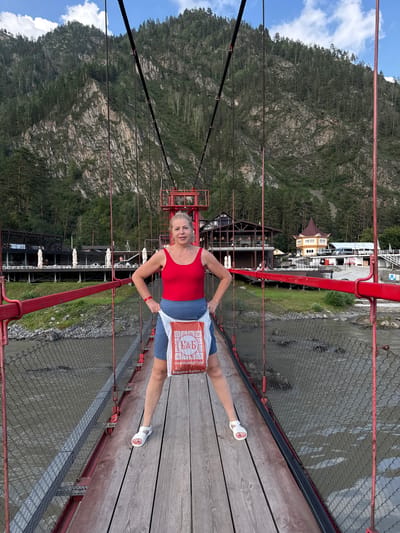 A blonde woman stands on a wooden suspension bridge in Barangol, Russia, holding a white plastic shopping bag. She is wearing a red tank top and blue shorts. The bridge spans a muddy river, with a chain-link fence along the sides. In the background, there are buildings with red accents and a steep, forested mountain. The sky is partly cloudy.
