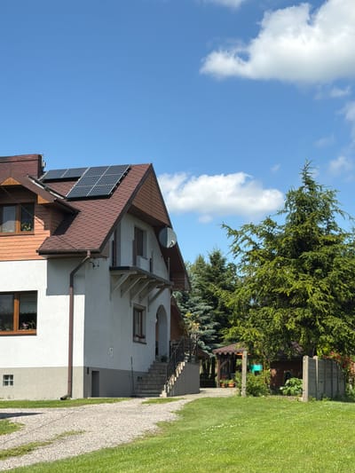 The image depicts a residential property in Strzyżowice, Poland, under a clear blue sky with scattered white clouds. The main subject is a two-story house with a brown shingled roof, featuring an attached section with light-colored siding. Solar panels are visible on the roof. The house has several windows, a brown downspout, and an arched entryway leading to a set of stairs. In the foreground, a gravel driveway curves into a well-maintained green lawn dotted with small white flowers. To the right of the house, there is a large evergreen tree, and behind it, a small wooden structure resembling a shed or gazebo with a brown roof, and a concrete fence. The overall scene suggests a sunny day in a semi-rural or suburban setting.