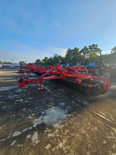 The image displays an outdoor scene featuring an array of large red agricultural implements, likely disc harrows or cultivators, parked on a concrete or paved lot in Karnobat, Bulgaria. The implements are prominently featured in the foreground and midground, showcasing their robust red frames and numerous discs. On the closest implement, the text "SDEI-K" is visible, while a second implement clearly displays "KAMIT" and "6011-30" in white lettering.

Several blue tractors are parked in the background, partially obscured by the red machinery. To the left, a line of vibrant green trees frames the horizon against a clear, bright blue sky, indicating sunny weather. The concrete ground exhibits weathering and some discoloration. In the distant background on the right, faint industrial-looking structures or power lines can be seen.

The bright sunlight suggests it is late morning or early afternoon. The scene depicts a static display or storage area for agricultural equipment, possibly at a dealership or a large farm. No people are visible, and no active work is taking place.