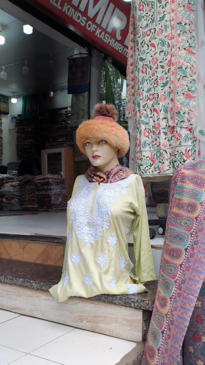 This is a medium-shot, eye-level, exterior photograph of a storefront in Dharamshala, India, featuring a mannequin dressed in traditional clothing. The mannequin, positioned on the right side of the frame, is wearing a pale yellow tunic with white floral embroidery on the neckline and sleeves, a colorful patterned scarf, and an orange, fur-trimmed hat. Behind the mannequin, on the right, is another mannequin or display draped with fabric featuring a large paisley pattern in shades of pink, teal, and yellow. To the left of the mannequin is a storefront with stacks of folded textiles and a red sign above that reads "ALL KINDS OF KASHMIRI ITEMS." The storefront appears to be on a street or walkway with tiled flooring in the foreground. The lighting suggests it is daytime, with some overhead artificial lights visible inside the shop. The overall scene depicts a retail environment specializing in Kashmiri goods.