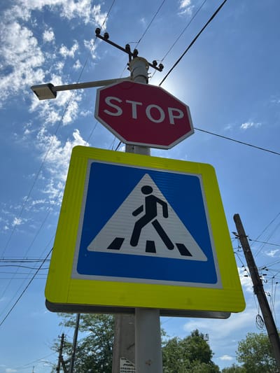 This low-angle, outdoor photograph captures a bright, sunny day with a clear blue sky dotted with wispy white clouds. The scene features two traffic signs mounted on poles, set against the sky and some overhead electrical wires.

The prominent red, octagonal STOP sign is positioned to the left, with the word "STOP" visible in white lettering. To its right is a blue and white pedestrian crossing sign, bordered by a bright yellow reflective frame. The pedestrian symbol depicts a black silhouette of a person walking across diagonal stripes.

Further details include the bottom of a street light hanging below the STOP sign and a snippet of a utility pole with multiple wires. On the pole holding the pedestrian crossing sign, there is a small sticker with visible Russian text that appears to be contact information for a taxi service: "TAXTA TAR «РУСЛАН» 8 (918) 036-11-0 8 (918) 429-42-1". The overall setting suggests an urban or suburban environment, likely in Russia, given the language on the sticker. The sun is bright, indicating it's likely midday. The signs suggest a road intersection or a pedestrian crossing area.