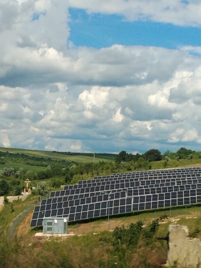 In the city of Budești, Moldova, an outdoor scene unfolds under a partially cloudy sky, featuring a sprawling solar panel installation. The foreground is dominated by rows of dark, rectangular solar panels, arranged on a grassy slope. To the left of the solar panels, a small, gray utility box with illuminated windows stands on a concrete base. Further up the slope, a small shrine with a cross and figurines is visible amidst greenery. The background transitions to rolling hills covered in lush vegetation and trees. The overall environment is rural and suggestive of renewable energy infrastructure.