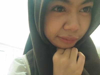 A close-up, low-angle, indoor selfie shows a young woman wearing a dark brown hijab and a white garment. Her face is illuminated by bright light, likely from windows in the background which are out of focus. Her right hand is supporting her chin. The scene appears to be in Tibojong, Indonesia, based on the metadata.