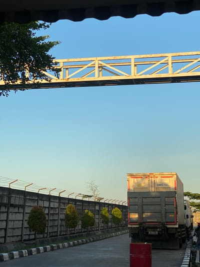 A daytime, exterior shot captures a scene in the vicinity of Gambut Barat, Indonesia. In the foreground, a red barrel sits on asphalt. Behind it, the rear of a white semi-trailer truck is visible, with its license plate reading "DA 8056 DA". The truck is parked on a paved surface adjacent to a concrete wall topped with barbed wire. A row of small, manicured evergreen trees lines the other side of the wall. Above the wall, a yellow metal overpass with diagonal bracing stretches across the frame against a clear blue sky. A portion of a leafy green tree is visible in the upper left corner, casting dappled shadows. The scene appears to be an industrial or transportation hub, possibly near a highway or freight terminal.