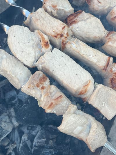 The image shows several skewers of raw meat, likely pork, being grilled over hot coals. The meat pieces are cut into uniform chunks and arranged tightly on metal skewers. Some pieces show slight browning on the edges, indicating the beginning of the cooking process. The background consists of dark, smoldering charcoal, with visible embers and smoke rising. The scene appears to be an outdoor barbecue. The image does not contain enough information to determine the specific location in the city of Oktyabrsky, Russia.