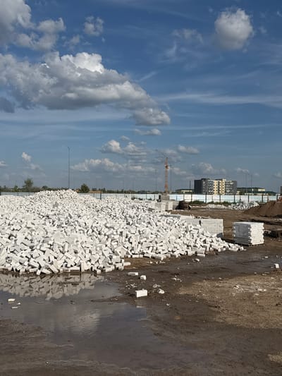 The image depicts a construction site in Zaşağan, Kazakhstan, on a bright, sunny day. The scene is dominated by a vast pile of white bricks, neatly stacked in some areas and more randomly strewn in others, suggesting ongoing construction work. Beyond the bricks, the ground appears to be a mix of mud and dirt, with a large puddle reflecting the sky, indicating recent rain or water usage.

In the background, a line of construction fencing separates the site from a cityscape. Several modern buildings, varying in height and architectural style, are visible, some appearing to be under construction themselves. A construction crane stands tall in the distance, a common sight at such locations. Streetlights dot the horizon, and scattered trees add a touch of greenery to the otherwise industrial landscape.

The sky above is a vibrant blue, dotted with fluffy white clouds and wispy cirrus formations, typical of a fair weather day. The lighting suggests it is daytime, likely late morning or early afternoon, given the bright sunlight casting shadows. There is no visible text in the image. The overall impression is one of active development and urban expansion.
