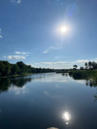 The image depicts a serene natural scene on a clear, sunny day. The sun is high in the sky, indicated by its bright glare and lens flare, suggesting it is midday. Scattered white clouds drift across a vivid blue sky, casting subtle shadows.

The foreground is dominated by a body of water, likely a river or lake, which acts as a mirror reflecting the sky, clouds, and the lush green foliage lining its banks. The water's surface is mostly calm, with ripples visible where the sunlight hits it. On the left side of the frame, a dense line of trees and other vegetation forms the shoreline, creating a natural boundary. These trees are full of green leaves, indicating a warm season.

The right side of the image shows more of the reflective water surface, with a partial view of what appears to be a wooden structure at the very edge. The composition is oriented vertically, as if the photo was taken looking up or down a waterway.

No people or specific objects are prominent, focusing the viewer's attention on the landscape itself. The overall mood is peaceful and tranquil, evoking a sense of natural beauty and stillness. The location is identified as Shyroke, Ukraine. There is no visible text in the image.
