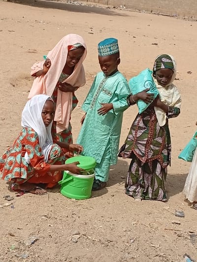 This outdoor photograph features four young children gathered on unpaved, sandy ground under bright daylight. A girl on the left, wearing a white headscarf and a patterned red, orange, and green dress, squats down, holding open a vibrant green portable cooler with a ribbed texture. Behind her, another girl in a light peach headscarf and a patterned dress, and a boy in a light green tunic (boubou) and a blue kufi (cap), lean in, looking intently into the cooler. On the right, a fourth girl, dressed in a patterned headscarf and long dress, stands holding a light blue plastic bag with an indistinct white logo or text.
The children appear to be engaged in an activity centered around the cooler, possibly sharing or preparing to distribute its contents, which are not clearly visible. Their expressions are focused and attentive. The scene is set on dry, sandy terrain, scattered with small debris, indicative of a warm, possibly arid environment. The bright sunlight casts strong shadows, suggesting it is midday or early afternoon. The traditional attire worn by the children and the natural surroundings align with the provided location context of Fanisau, Nigeria. Parts of another child in a white dress are visible on the far right.