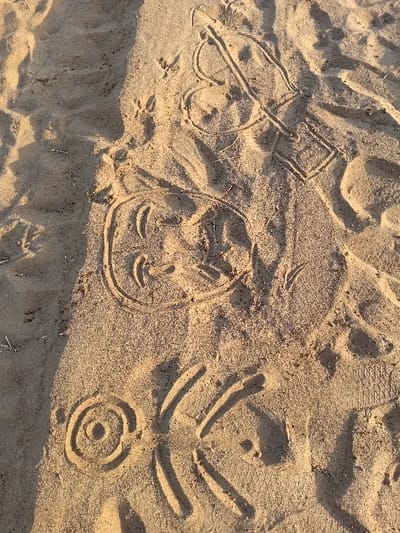 The image depicts sand art of varying complexity. A spade symbol is drawn with a square-shaped top. A simple cartoon face features large eyes and a small, curved smile. A circular target-like design has concentric circles and several lines radiating outwards. The composition includes small footprints, suggesting animal activity. The sandy surface is textured and lit with a warm tone, indicating sunlight.