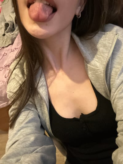 The image depicts a close-up, angled view of the upper torso and head of an unidentified individual located indoors in Волховец, Russia. The subject, likely female, has long, dark brown hair and is seen sticking their tongue out of an open mouth. A small, silver-colored hoop earring is visible in the subject's right ear. The individual is wearing a light grey, hooded, zipper jacket, partially unzipped, over a black, ribbed tank top with a two-button placket. In the background, to the upper left, a light pink or purple patterned fabric with white script or designs is visible, likely a blanket or sheet. Below this, a darker, indistinct surface is present. The lighting is consistent with an indoor setting.