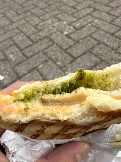 A partially eaten panini, filled with pesto and melted cheese, is held by a hand. The sandwich is wrapped in white paper. The background consists of grey paving stones laid in a herringbone pattern. The location cannot be determined from the image.