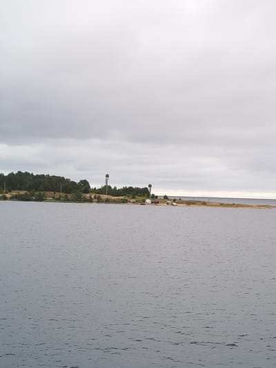 A tranquil, overcast day unfolds over a body of water in Shal'skii, Russia. The vast expanse of grey water stretches towards a low-lying, tree-covered shoreline. On the shore, several structures are visible: two prominent water towers, one closer than the other, stand tall against the cloudy sky. Near the water towers, a cluster of white tents and several vehicles suggest a temporary encampment or recreational area. The shoreline is a mix of sand and vegetation, with sparse trees dotting the landscape. A person is faintly visible walking on the far right of the shore. The overall atmosphere is one of quiet serenity, with the muted colors of the sky and water reflecting the calm weather.