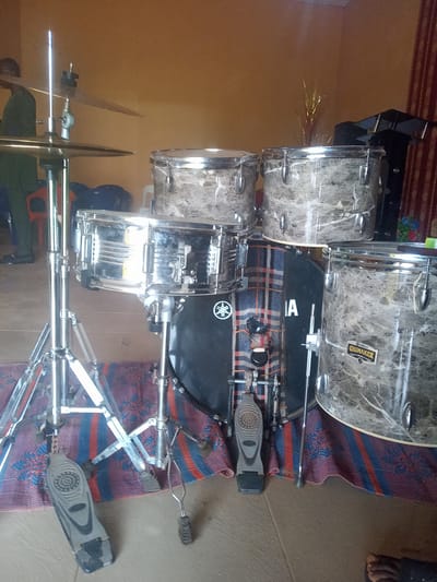 A drum set, consisting of a black bass drum with "YAMAHA" logo, a chrome snare drum, three marble-patterned toms, a cymbal stand with cymbal, and bass drum pedals, is positioned on a multicolored patterned rug. The backdrop is a yellow wall and red and maroon curtains. The scene appears to be indoors. The location is Nkwelle Ezunaka, Nigeria. An individual wearing green attire is partially visible on the left side.