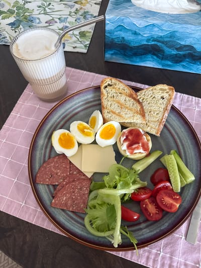 A close-up overhead shot shows a breakfast spread on a dark wood table, covered partly by a pink checkered placemat. A tall, fluted glass filled with a frothy beverage, likely coffee with milk, is positioned on the upper left, with a metal straw. To the right of the drink is a tissue box with a blue wavy pattern.
The main subject is a large, dark-rimmed plate filled with various food items. These include:
* Two slices of toasted bread, one angled on top of the other.
* Four halves of hard-boiled eggs, with visible yolks.
* A slice of pale yellow cheese.
* Several thin slices of salami with visible pepper flecks.
* A small, round pastry or cracker topped with white cream and a swirl of red jam.
* A small pile of green lettuce.
* Several cherry tomatoes, some halved, showing their red interiors.
* Sticks of sliced cucumber.
The scene is devoid of any people and the setting does not provide specific visual cues to pinpoint its location within Gatineau, Canada. The lighting appears to be natural, casting soft shadows.