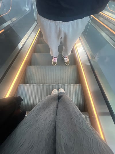 The image provides a high-angle view looking down an escalator in an indoor, modern architectural setting within the city of León, Mexico. Two individuals are visible. In the immediate foreground, the lower torso and legs of a person are shown, wearing light grey textured wide-leg pants and light-colored sneakers; a dark object is partially visible on the far left. One step below, another individual's lower torso and legs are visible, dressed in light grey sweatpants and two-toned sneakers, one with pink laces and the other with white or cream laces, both featuring green/earth-toned bases. The escalator steps are metallic and ribbed. Transparent glass balustrades flank the steps, with metallic handrails above them. A warm orange-yellow LED light strip runs along the base of the handrail on both sides of the escalator. Both subjects are standing stationary on the moving escalator.