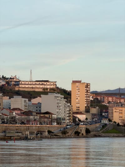 This image depicts a panoramic view of the city of Peso da Régua, Portugal, primarily focused on the urban landscape along the Douro River.

The foreground is dominated by the wide, placid waters of the Douro River, exhibiting ripples and reflecting the light sky. A red cylindrical buoy is visible near the left bank. On the river's edge, a concrete retaining wall supports a promenade and road. Along this waterfront, a small pier or dock structure with a light-colored building is present.

The midground features a dense urban area. A multi-lane road runs parallel to the river, with numerous parked vehicles, predominantly cars, lining its edges. Behind this road, a variety of buildings are observed: some appear as older, multi-story residences with pitched roofs, while others are modern, rectangular structures. A prominent light beige, multi-story building, appearing residential, stands near the right side of the frame with "Régua Douro" visible on its facade.

The background consists of steep hills rising behind the urban sprawl. These hills are largely covered with buildings, including a long, white or light-colored multi-story structure with numerous windows, situated high on the slope. Further up the hill, a tall, grey lattice transmission tower is visible. To the far right, a very tall, slender, light brown residential or commercial building with numerous small windows dominates a section of the skyline. In the distant background, a multi-lane concrete bridge structure is visible, spanning across a valley or gorge. The sky above is a pale blue with faint, wispy cloud formations. No individual people are distinctly visible, but their presence is implied by the urban environment and vehicles.
