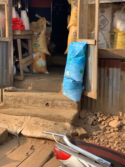 This image depicts the entrance of a commercial establishment, likely an agricultural or animal feed supply store, located in Kabba, Nigeria. The scene is illuminated by strong daylight, indicating an outdoor setting.
The foreground features rough wooden planks on the ground, with a partial view of a silver motorcycle or scooter in the lower right. The motorcycle shows a black and red striped seat, a silver grab rail, and a red taillight. Beyond the planks, the ground is unpaved, consisting of dry earth and scattered small rocks.
Two unadorned concrete steps lead up to an open doorway. The shop's facade consists of wooden framing and corrugated metal sheeting on the right. Leaning against this structure, to the right of the steps, are two stacked, light blue plastic sacks marked "BLUECROWN PREMIUM FISH FEED 15KG," each featuring a fish graphic and a crown logo.
Inside the doorway, various sacks are visible. In the center, a large tan sack displays "Grand Cereals Limited" text and a chicken graphic. Directly below it, another sack features a prominent image of a white chicken. To the far left, within the shop's opening, multiple white plastic poultry feeders and several red plastic components (possibly feeder bases or waterer parts) are stacked on or near a wooden frame. The interior of the shop beyond these items appears dark. No individuals are visible in the frame.