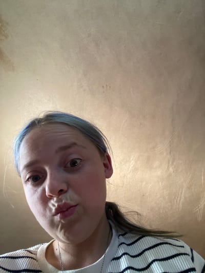 The media file is a close-up, downward-facing selfie of a young person in Nova Praha, Ukraine. The subject has fair skin, brown eyes, and blue-tinted hair. They are wearing a white and black striped sweater and a silver necklace. The person's mouth is slightly open, and they appear to be making a playful or curious expression. The background is a textured, light-brown surface, possibly a wall or ceiling, with a subtle sheen. There are no clear indicators of the time of day, weather, or specific location beyond the provided context of Nova Praha. The overall mood is informal and candid.