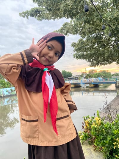 A young female subject is depicted outdoors in Kalitengah, Indonesia, facing the camera. She wears a light brown uniform shirt with dark brown collar and cuff accents, a dark brown skirt, and a dark brown hijab. A red and white neckerchief, secured with a green slide, is worn around her neck. Her left hand is raised to her temple with fingers spread in a gesture. The background features a narrow waterway, spanned by a multi-colored pedestrian bridge (blue, yellow, green sections) in the mid-ground. Distant low-rise structures and vegetation are visible on the far bank. The sky is overcast, displaying a subtle orange tint near the horizon. Overhead, a large tree with light green and white variegated leaves supports a string of black-housed lights. A concrete embankment runs along the immediate foreground, adjacent to a bush bearing yellow flowers.