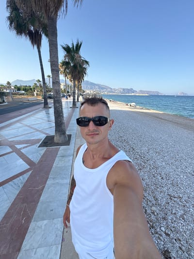 A selfie is taken by a man wearing a white tank top and black sunglasses, standing on a pebble beach. He is looking at the camera with a neutral expression. Behind him, a paved walkway with a marble pattern lines the beach, with palm trees and a street visible on the left. The sea is to his right, with a distant coastline and mountains in the background. The scene appears to be in Altea, Spain, known for its picturesque beaches and Mediterranean views. The sky is clear and blue, suggesting a sunny day.