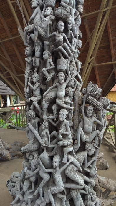 A large, intricately carved wooden sculpture, appearing to be a single piece of light-colored, weathered wood, is centrally positioned. The sculpture is densely populated with numerous human figures, both male and female, depicted in various active poses: climbing, reaching, carrying, and sitting. Many figures bear baskets, bundles of goods, and containers on their heads and shoulders. The figures exhibit stylized features and minimal clothing. The sculpture rests on an earthen or compacted ground surface. Overhead, a wide, multi-faceted wooden roof structure is visible, supported by prominent timber beams, suggesting an open-air pavilion. In the background, beyond a low wooden railing, lush green vegetation, including plants with red flowers, is present. Portions of other buildings with brown tiled or wooden roofs are visible to the left and right. This scene is located at the Cultural Heritage Centre in Arusha, Tanzania.