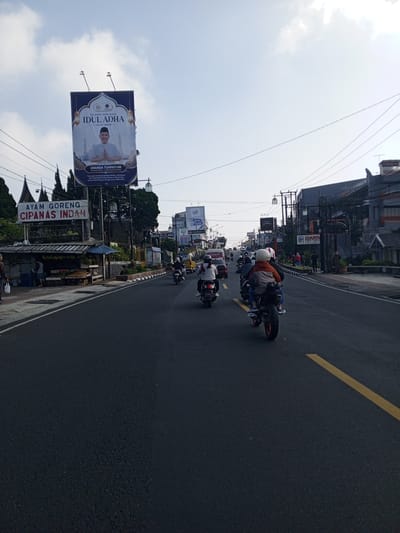 This image depicts a street scene in the city of Weddagala, Sri Lanka, captured during daylight hours under a bright, partly cloudy sky. The perspective is from a low angle, looking forward along a wide asphalt road marked with a solid yellow line on the right edge of the visible lane.
On the left side of the road, a large billboard is mounted on a tall pole. The billboard features the Indonesian text "SELAMAT HARI RAYA IDUL ADHA 1447 H / 2026 M" (Happy Eid al-Adha 1447 H / 2026 AD) and displays a portrait of a smiling man. Below the portrait, the text identifies him as "ANANDA TOHPATI NR, ANGGOTA DPR RI - FRAKSI PARTAI NASDEM" (Member of the House of Representatives - Nasdem Party Faction) and includes an Indonesian quote: "Semoga semangat pengorbanan, keikhlasan dan kepedulian terus tumbuh di hati kita untuk membangun bangsa yang lebih baik." (May the spirit of sacrifice, sincerity, and care continue to grow in our hearts to build a better nation). Beneath the billboard, a roadside stall with a corrugated metal roof is present, displaying a sign with Indonesian text "AYAM GORENG CIPANAS INDAH" (Fried Chicken Cipanas Indah). Produce, including bananas, is visible at the stall. Two individuals are near this stall, one partially obscured by the foreground. Coniferous trees and numerous utility lines on poles are visible in the background on the left.
On the road, multiple motorcycles are traveling in the same direction as the camera. Closest to the viewer, a black motorcycle with a red rear wheel carries two riders; the front rider wears an orange/brown jacket and a white helmet, and the passenger wears blue jeans. Ahead, another person in a dark helmet and light shirt rides a separate motorcycle. Additional motorcycles and a red car are visible further down the road.
The right side of the road features a paved sidewalk with a curb, some vegetation, and multi-story buildings. Several signs are visible on this side, including "STIEBIS 69" on a dark building, "IDAMA" on a lower sign, and a brown sign for "BIARA S. CLARA" (Convent of St. Clara). A more distant billboard displays the word "CLASSY" and numerical information "50 ribu 39,000". A group of pedestrians, some dressed in red, are walking on the right sidewalk in the mid-ground. The scene depicts an active urban or semi-urban environment with ongoing vehicle traffic and commercial establishments.