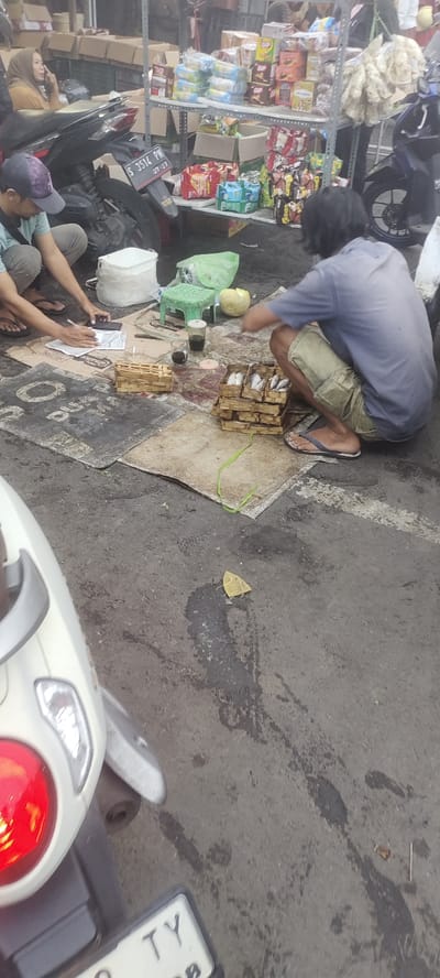 The scene takes place on a street in Mojokerto, Indonesia, at the coordinates -7.468 latitude and 112.438 longitude. A man wearing a blue shirt and khaki shorts is crouched on a rug, sorting through a pile of small, woven baskets containing fish. Another man in a blue shirt and beige pants is seated on the ground in front of a stack of cardboard boxes, writing on a piece of paper. A white plastic container sits nearby, along with a green stool, a small, green bag, and a melon. There is a motorcycle in the background, with its license plate visible. The man crouched on the rug is facing away from the camera, while the other man is facing toward the camera. The rug is spread out on the street, and the surrounding area is paved with asphalt. A white motorcycle is parked nearby, with its taillight visible in the bottom left corner of the image.
