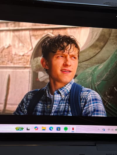 This media file is a photograph of a digital screen displaying a scene from a movie or TV show, captured in Bar Gada, Nigeria. The image is rotated 90 degrees clockwise.
The central subject on the screen is a young man, likely Tom Holland, with wet, curly brown hair. He is looking upwards and slightly to the side with his mouth slightly open, suggesting an expression of surprise, action, or mild distress. He is wearing a blue and white plaid shirt and a blue backpack. The background of the movie scene features old, weathered architectural elements, including large stone or concrete arches and walls in muted tones of green, grey, and pale brown.
To the right side of the screen's content, a vertical Windows taskbar is visible. It displays several icons, including those for Wi-Fi, volume, Netflix, Spotify, Microsoft Edge, File Explorer, a multi-colored square icon (possibly Teams or Paint 3D), and a distinct green and blue application icon at the bottom. The border of the screen, or bezel, is black, and part of a label, "BANG & O", is visible on the right side of the device, likely indicating the manufacturer (Bang & Olufsen). The screen surface itself shows some streaks and smudges, suggesting it has been used or handled.
The overall image captures the moment a person is watching content on a digital device, with elements of both the content and the device's interface and physical form visible.