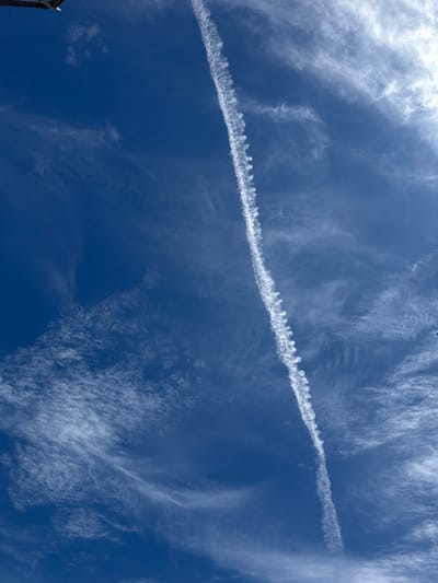 A photograph taken from a low angle looking up at a bright blue sky with wispy white clouds. A long, thin contrail from an aircraft cuts diagonally across the frame from the upper left to the lower center, dissipating into smaller cloud fragments. There are hints of a dark object, possibly part of a structure or a bird, in the upper left corner of the image. The overall impression is of a clear day with some atmospheric activity. This image was taken in the Borough of Swale, United Kingdom.