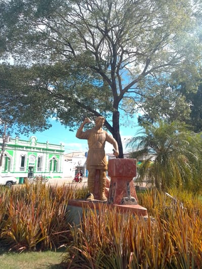 A bronze statue of a blacksmith is centrally located in an outdoor public space, likely a plaza or park, in Cáceres, Brazil. The statue depicts a man with a hammer raised over an anvil, engaged in the act of forging. He is wearing a textured garment that suggests work clothes. The statue stands on a multi-tiered, rough-hewn base that is reddish-brown in color.

In the background, a large, mature tree with dense green foliage dominates the upper portion of the image. Sunlight filters through its branches, creating dappled light effects. To the right of the statue, a palm tree with vibrant green fronds adds to the tropical atmosphere.

Beyond the statue and trees, a two-story building with a white facade and bright green trim is visible. It features arched windows and a balcony on the upper level, suggesting it is a historic or significant structure, possibly a government building or commercial establishment. Several bicycles are parked in front of this building, and further to the right, a motorcycle with two occupants is in motion. A white vehicle is partially visible on the left side of the image.

The foreground is filled with dense, dry-looking grasses with reddish-brown and yellow hues, creating a textured border around the statue. A small patch of green grass is visible in the lower left corner. The sky is a clear, bright blue with a few wispy white clouds. The overall scene conveys a sense of community and local culture, highlighting a historical or artisanal element within the urban environment of Cáceres.