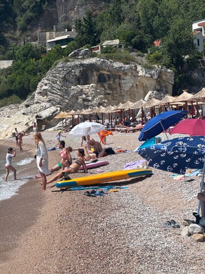 The image depicts a lively beach scene in Mirište, Montenegro, characterized by a rocky shoreline and a multitude of beachgoers enjoying the sun and sea. The beach itself is composed of small pebbles, with the water gently lapping at the shore. Several people are visible in and near the water, with some wading in and others relaxing on the sand. Beach umbrellas, both straw and fabric, provide shade for those lounging on the beach. Various beach accessories, including towels, inflatable toys, and surfboards, are scattered across the sand. In the background, a steep, rocky cliff face rises behind the beach, with lush green trees and a few buildings dotting the hillside, suggesting a coastal settlement. The overall atmosphere is one of leisure and recreation on a bright, sunny day.