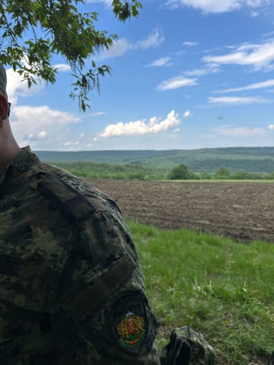 The image shows a field in Kostena Reka, Bulgaria. A person wearing a camouflage uniform with a Bulgarian emblem on the sleeve is partially visible in the foreground. The field is bordered by grass and a line of trees leading to a forested area on the horizon. The sky is blue with scattered clouds.