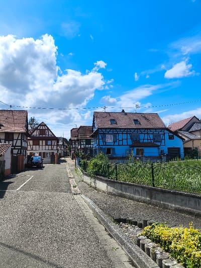 The image depicts a residential street scene in Oberschaeffolsheim, France, under a clear blue sky with scattered white cumulus clouds. The perspective is from ground level, looking down a paved street with a slight incline.
On the left side of the street, multiple traditional half-timbered houses are visible. The first house on the immediate left is partially obscured, showing a brown-tiled roof and white facade with dark timbering. Further down the street, a dark-colored sedan is parked in a marked parking space on the left. Beyond the parked vehicle, additional half-timbered houses with brown-tiled roofs line the street, characterized by white facades and dark brown timber frames.
On the right side of the street, a prominent two-story house with a blue facade and dark brown half-timbering is visible. This house features multiple windows, some with light-colored shutters, and a brown-tiled roof with two visible skylights and roof antennae. In front of this house, a low concrete wall supports a green metal fence, behind which overgrown vegetation is present. Closer to the foreground on the right, a small patch of yellow-green foliage is bordered by cylindrical concrete elements. The street itself is composed of a light-colored aggregate material, with a narrow concrete curb separating it from a gravel-lined gutter and a narrow, raised sidewalk on the right. Utility wires are visible spanning across the upper portion of the frame, above the houses. No individuals are present in the scene, and no specific actions or interactions are depicted.