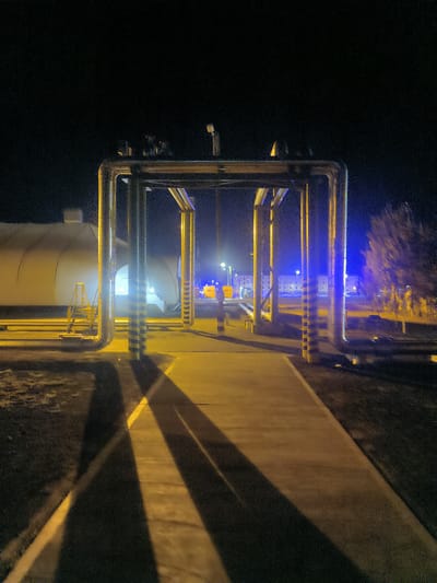 The image depicts an outdoor industrial or utility area at night in Қосшағыл ауылдық округі, Kazakhstan. Prominently featured is a large metal pipe structure, illuminated by yellow and blue lights, casting long shadows across a concrete pathway. The pipes are supported by industrial-style pillars, some of which have black and yellow striped safety markings. To the left, a large, white, tent-like structure is visible, with a yellow metal staircase leading up to it. In the background, there are buildings and streetlights, suggesting a populated or functional area. A tree with sparse foliage is visible on the right, illuminated by blue light. The overall atmosphere is industrial and somewhat stark, illuminated by artificial lighting against a dark night sky.