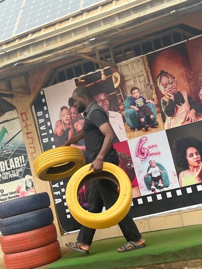 The image shows a man holding a yellow tire, standing in front of a wall covered with large posters. The posters feature images of people, including children and women, with some text elements. One poster appears to be a birthday announcement with a balloon and the text "HAPPY BIRTHDAY." Another poster displays the name "DAVID." Below the main posters, there are more posters with text. Some of the visible text includes "TECK LIMITED," "OLAR!! A POINT TO SAVE THIS PERIOD," and "Plaza Saburi 1, Mopol Barracks." Several other tires, colored blue and red, are stacked on the ground. The setting appears to be outdoors or in a semi-covered area, with a glimpse of solar panels on the left side of the image and a grassy surface on the right. The man is wearing dark pants and sandals. The overall impression is of a vibrant, possibly commercial or community space, decorated with photographic art and repurposed tires. The lighting suggests it is daytime.