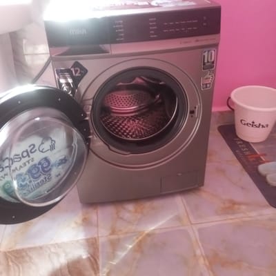 This image depicts an indoor scene within the city of Mahiakalo ward, Kenya. The primary subject is a light grey/silver Mika brand front-loading washing machine, positioned centrally on a tiled floor. The washing machine's circular door is open, extending to the left, revealing an empty, perforated metallic drum inside. Visible labels on the machine indicate a "12 kg" capacity, "BRUSHLESS INVERTER MOTOR," "10 YEARS" warranty, and "MIKA CARE." The dark control panel above the drum features various wash cycle options, including "COTTON," "SPORTS," "QUICK WASH," "DELICATE," and "STEAM" technology.
To the right of the washing machine, a white plastic bucket with the word "Geisha" printed in black script is visible. This bucket rests on a dark-colored rectangular floor mat, partially visible, which bears white text, including "FRESH" and "KITCHEN." Light-colored footwear, possibly sandals or slippers, are partially visible on the mat, situated to the right of the bucket. The background features a solid pink-painted wall. The floor consists of light-colored, possibly beige or pale marble-patterned, square tiles. Bright illumination from an unseen source creates reflections on the washing machine's surface and the floor. No individuals are present in the scene, and no specific actions or interactions are depicted.