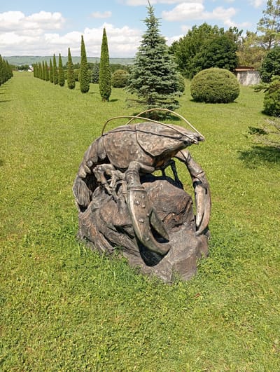 This media file is a photograph of a bronze sculpture of a lobster, lying on a grassy knoll in an outdoor setting. The sculpture is detailed, with a textured, weathered appearance, and it appears to be made of dark metal. The lobster sculpture is the primary subject, with its claws and segmented body clearly visible.
The setting is a well-maintained park or garden. The ground is covered in lush green grass, with some scattered small yellow flowers. In the background, there are various trees and bushes, including manicured conical evergreen shrubs planted in a line, receding into the distance. Further back, a row of larger trees and a rounded, sculpted bush are visible. The sky is a clear blue with fluffy white clouds, suggesting a pleasant day. The lighting indicates it is daytime, likely late morning or afternoon, with sunlight casting shadows.
No people are visible in the image. The activity taking place is simply the display of the sculpture in its environment. There are no visible text elements within the image. The overall impression is one of a peaceful, scenic outdoor location where art is displayed. The location context of Otradnaya, Russia, provides a geographical reference for the scene.