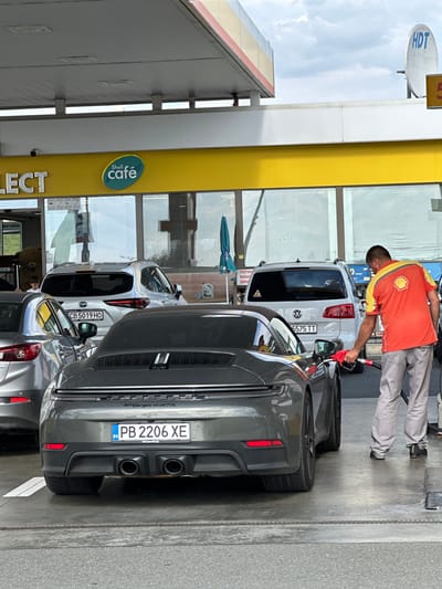 A gas station attendant in an orange and yellow Shell uniform dispenses fuel into a dark gray Porsche 911 Carrera GTS at a Shell gas station. The Porsche has a Bulgarian license plate reading PB 2206 XE. To the left of the Porsche is a silver Mazda CX-5. In the background, a white Volkswagen Touran SUV is parked, also at a gas pump. The gas station features a yellow canopy with "Shell café" signage. The scene appears to be in the city of Drazhevo, Bulgaria, as indicated by the license plate and local signage. The sky is partly cloudy.