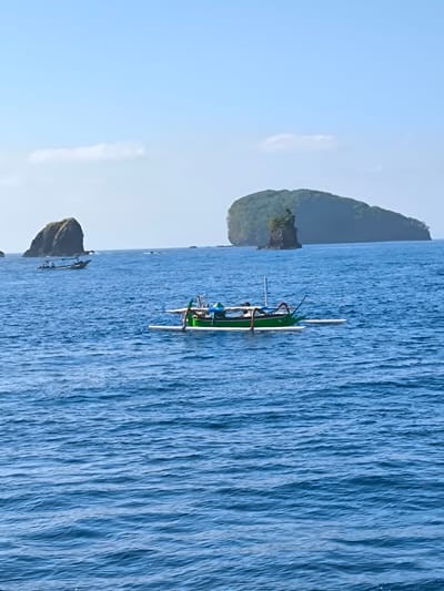 A vertical, slightly grainy photograph captures a serene ocean scene with two boats and a distant island. In the foreground, a green and white outrigger canoe with an awning is anchored in calm, blue water. The canoe appears to be occupied by at least two people, though their details are indistinct. In the mid-ground, a larger, lighter-colored boat with passengers is visible moving away from a rocky outcrop. Further in the distance, a large, lush island with a prominent rock formation in front of it dominates the horizon. The sky is clear and pale blue with a single wispy cloud. The location is the ocean off the coast of Karangasem, Indonesia, a region known for its beautiful islands and clear waters.