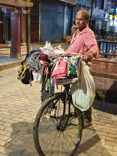 A man in a pink shirt and green pants stands next to a bicycle loaded with colorful textiles. The bicycle has a front rack with fabric bundles, and a white plastic bag hangs from the handlebars. The man holds the bicycle's handlebars. In the background, there are closed shop shutters and a brick wall. The ground is paved with bricks, and a shadow of the man and bicycle is cast on the ground. The scene appears to be a street in Kathmandu, Nepal, possibly at night or in the early evening due to the lighting.