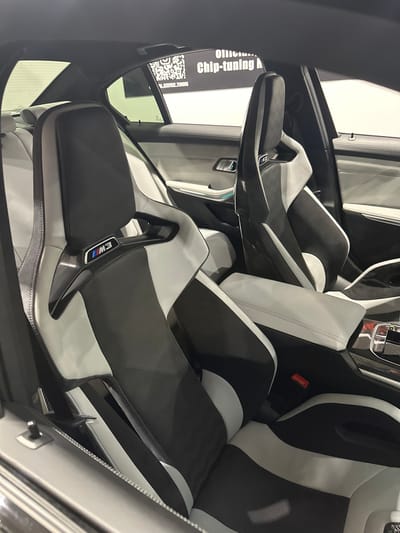The image displays the interior of a high-performance BMW M3 vehicle, specifically the front and partially visible rear passenger compartments, located in Wolica, Poland. The perspective is from the rear-right of the cabin, looking forward. Two racing-style front bucket seats dominate the foreground and midground. Both seats feature a distinct black and light grey/white upholstery pattern, incorporating perforated textile, smooth leather-like material, and glossy black carbon fiber trim, particularly around the seatbacks and bolsters. Each front seatback prominently displays a metallic BMW M3 emblem with the characteristic M colors. Between the front seats, a light grey upholstered center armrest is visible, with a red seatbelt buckle situated near the base of the passenger seat. The rear seats and interior door panels are upholstered in a light grey/white material. A metallic door handle and a subtle light-blue accent line are visible on the rear-left door panel. Through the rear windows, a black sticker is affixed, displaying a white QR code and text fragments such as "Official," "Chip-tuning," and an Instagram handle "_M_SERVICE_TUNING." The interior appears to be illuminated by artificial light, suggesting an indoor setting. No individuals are present in the vehicle.