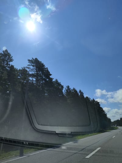 The image shows a view from inside a vehicle, looking out through the windshield. The sun is bright and high in a clear blue sky with some wispy clouds. A line of dark green pine trees runs along the left side of a paved road, which curves to the right and recedes into the distance. The road has white lane markings. In the distance, there appear to be a few vehicles on the road. The foreground of the image is dominated by the dark interior of the vehicle, likely the dashboard and windshield frame. The overall scene suggests a sunny day driving through a wooded area. The location is identified as Jūraskalni, Latvia.