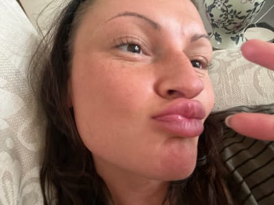 A close-up selfie of a woman with brown hair, light skin, and brown eyes. She is making a kissing face with pursed lips. A finger is partially visible in the lower right corner. The background includes a patterned white surface, possibly a cushion or upholstery, and a floral patterned vase. The location is undetermined; there's no clear evidence indicating Tunbridge Wells, United Kingdom.