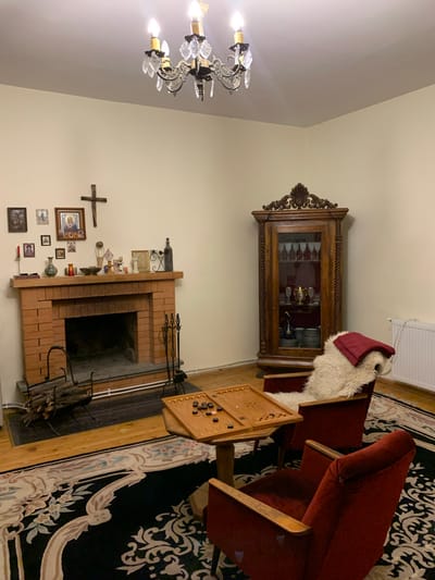 The image depicts a dimly lit, traditional living room setting. A red brick fireplace with a hearth containing logs and fireplace tools is the focal point on the left. Above the fireplace, a collection of religious icons and framed pictures adorn the wall, alongside a large wooden cross. A wooden mantelpiece displays various decorative items, including vases and candles.
To the right of the fireplace, a vintage wooden cabinet with glass doors showcases glassware and plates. In the foreground, two red velvet armchairs are placed around a wooden table with a backgammon board set up, suggesting a game in progress. A fluffy white sheepskin throw and a red blanket are draped over one of the armchairs. The room features a large, ornate black rug with a patterned border covering most of the wooden floor. A chandelier hangs from the ceiling, illuminating the space. The overall ambiance suggests a cozy, perhaps elderly, dwelling. There are no people present in the image. The scene is set in Mepiskalaki, Georgia.